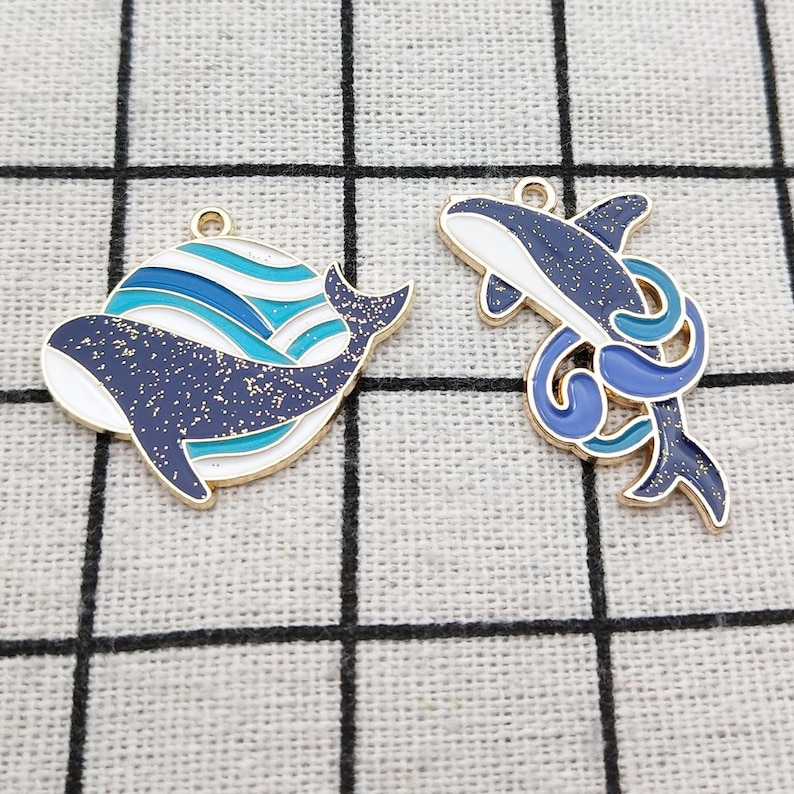 May include: Two gold-tone metal whale charms. One charm features a blue and white whale with waves, the other charm features a blue whale with a blue tail fin.