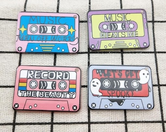 May include: Four colourful cassette tape pins with different designs. The first pin is pink and blue with the text "MUSIC TURN OFF WORRIES". The second pin is purple and yellow with the text "MUSIC HEAL SME". The third pin is pink and yellow with the text "RECORD THE BEAUTY". The fourth pin is blue and red with the text "LET'S GET SPOOKY" and two ghost figures.