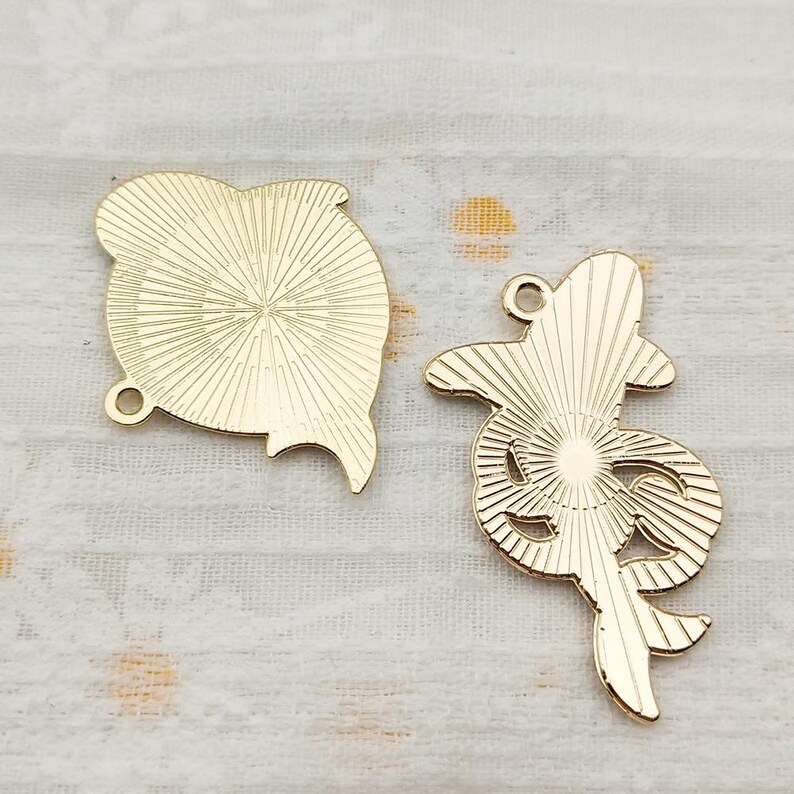 May include: Two gold-colored metal charms, one shaped like a heart and the other shaped like a stylized flower. Both charms have a textured surface with a sunburst pattern.