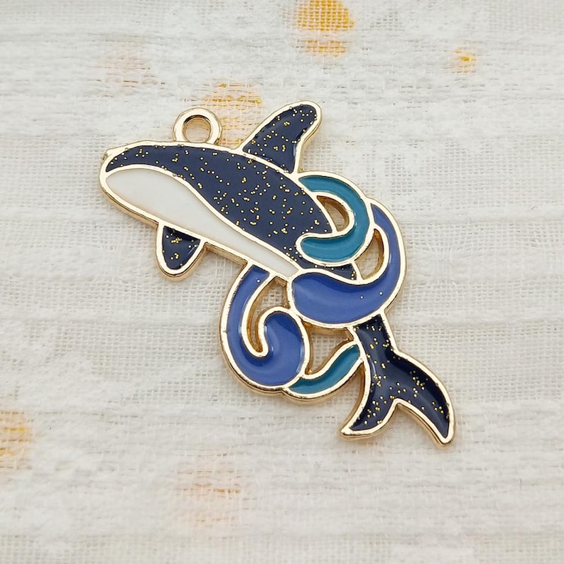 May include: Gold-tone metal charm featuring a blue and white orca with a blue and green swirl design. The orca has a white belly and a black back with gold glitter.