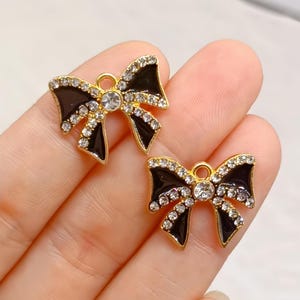 Peut inclure: Deux breloques en forme de nœud avec une finition en émail noir brillant, des bords en métal doré et des strass transparents le long des bordures et au centre. Chaque breloque comprend un petit anneau circulaire en haut pour la fixation.