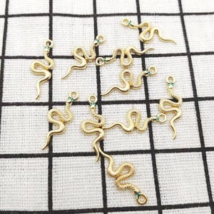 May include: A collection of gold-toned metal snake charms, each featuring a textured, winding body and two small green inlaid stones representing eyes. Each charm includes a small circular loop at the top for attachment to jewellery.