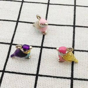 Könnte beinhalten: Drei kleine Vogel-Anhänger mit goldenen Akzenten. Die Vögel sind in verschiedenen Farben bemalt: lila, rosa und gelb.