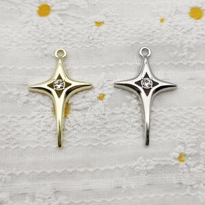 May include: Two four-pointed star-shaped pendants, one in a gold-tone finish and one in a silver-tone finish. Each pendant features a central clear round rhinestone set within a dark, diamond-shaped inlay at the centre of the star design.