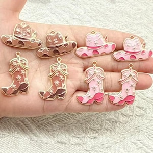 May include: A collection of eight metal charms shaped like cowboy hats and cowboy boots. The charms feature enamel detailing in shades of brown and pink, with white floral patterns and small metallic loops at the top for attachment.
