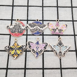 Puede incluir: Seis dijes de metal diferentes con diseños coloridos de mariposas y polillas. Cada dije tiene un acabado dorado y presenta un patrón en blanco y negro con una variedad de colores, incluyendo rosa, azul, morado y amarillo.