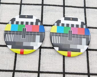 May include: Two circular pendants featuring a black and white test pattern design with a hole at the top. The design incorporates colourful bars and lines, with a yellow, red, and black border. Each pendant is approximately 3 cm in diameter.