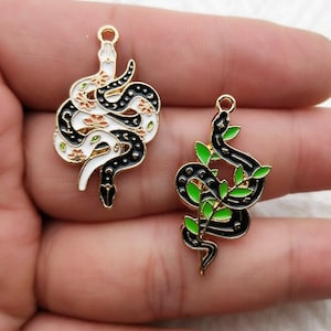 May include: Two gold-toned snake charms. One charm is black and white with floral accents. The other is black with green leaf accents. Both charms have a loop at the top for attaching to jewelry.