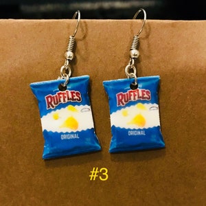 May include: A pair of silver dangle earrings featuring a blue and white Ruffle's potato chip bag charm. The bag has the text "Ruffles" and "Original" printed on it.
