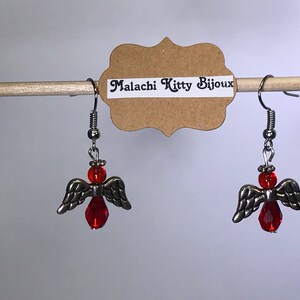May include: A pair of silver angel wing earrings with red glass beads. The earrings are hanging from a wooden dowel rod with a brown paper tag that reads "Malachi Kitty Bijoux".
