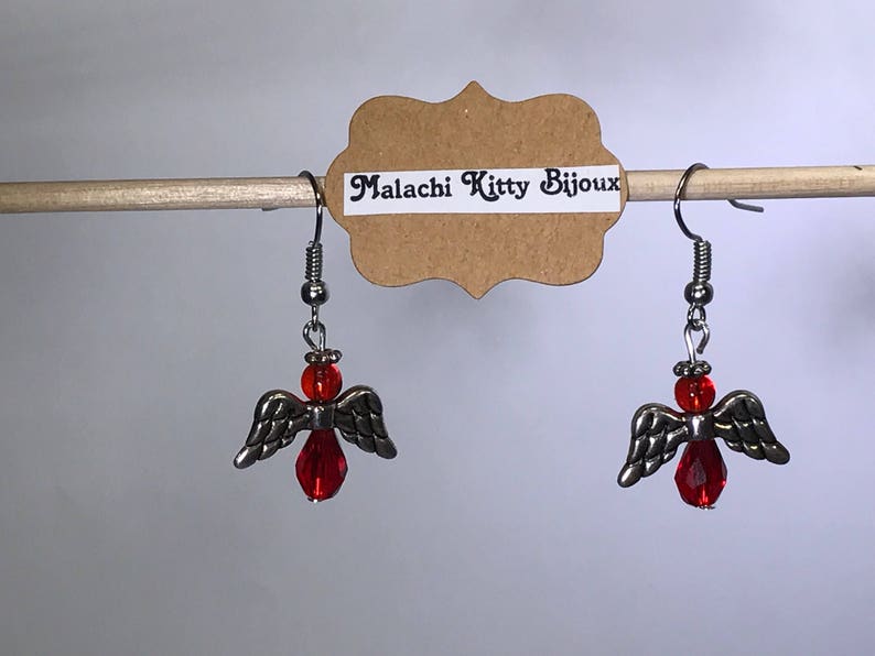 May include: A pair of silver angel wing earrings with red glass beads. The earrings are hanging from a wooden dowel rod with a brown paper tag that reads "Malachi Kitty Bijoux".