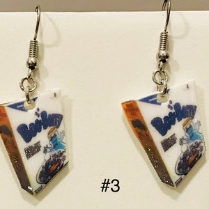 May include: A pair of silver dangle earrings with a white, blue, and orange cereal box charm. The box has the words "Boo Berry" printed on it. The charm is shaped like a triangle and has a glittery finish. #3