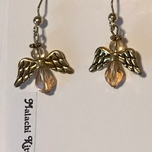 May include: A pair of gold angel wing earrings with clear crystal drops. The earrings are on a white card with the text "Fashion Jewellery" and "Malachi Kitty Bijoux".