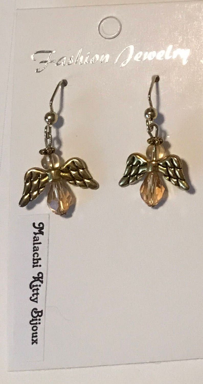 May include: A pair of gold angel wing earrings with clear crystal drops. The earrings are on a white card with the text "Fashion Jewellery" and "Malachi Kitty Bijoux".