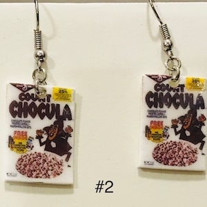 May include: A pair of silver earrings with a Count Chocula cereal box charm. The box is white with a red and brown illustration of the Count Chocula character. The text on the box reads "Count Chocula" and "Chocolate Flavored Marshmallow Bits".