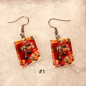May include: A pair of dangle earrings featuring miniature cereal boxes. The boxes are red and white with the text "Kellogg's Froot Loops" printed on them. The earrings are silver.
