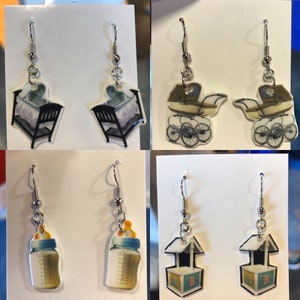 May include: Four pairs of baby-themed earrings. The first pair features a crib with a baby inside. The second pair features a baby carriage. The third pair features a baby bottle. The fourth pair features a toy block with the letters A and B.