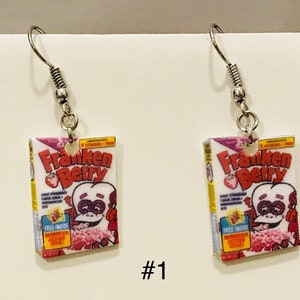 May include: A pair of silver earrings featuring a miniature replica of a Franken Berry cereal box. The box is white with red, purple, and yellow accents. The front of the box features a cartoon monster with a red strawberry on its head. The text "Franken Berry" is printed in red and white.