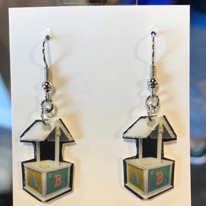 May include: A pair of silver dangle earrings with a white, yellow, blue, and black printed design of a toy block with a roof. The block has the letters A and B on it.