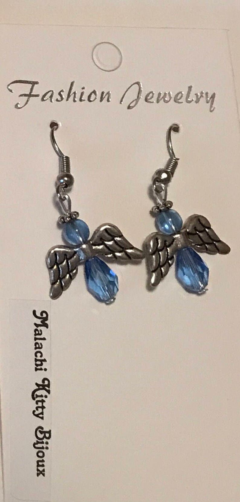 May include: A pair of silver angel wing earrings with light blue crystal beads. The earrings are on a white card with the text "Fashion Jewelry" and "Malachi Kitty Bijoux".