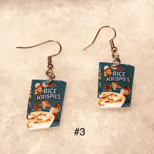 May include: A pair of silver dangle earrings featuring miniature replicas of Rice Krispies cereal boxes. The boxes are blue and white with the Kellogg's logo and the words "Rice Krispies" printed on them.