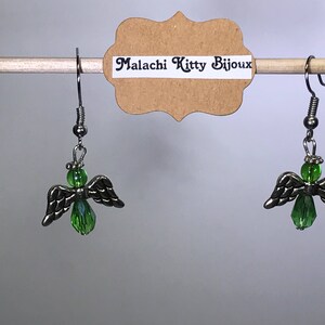 May include: A pair of silver angel wing earrings with green crystal accents. The earrings are hanging from a wooden dowel rod with a brown paper tag that reads "Malachi Kitty Bijoux".