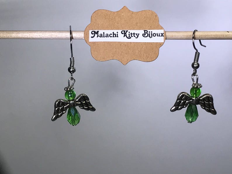 May include: A pair of silver angel wing earrings with green crystal accents. The earrings are hanging from a wooden dowel rod with a brown paper tag that reads "Malachi Kitty Bijoux".