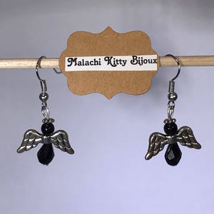 May include: A pair of silver angel wing earrings with black crystal accents. The earrings are hanging from a wooden dowel rod with a brown paper tag that reads "Malachi Kitty Bijoux".