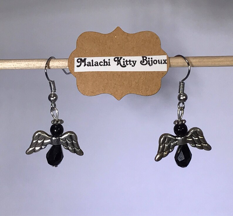 May include: A pair of silver angel wing earrings with black crystal accents. The earrings are hanging from a wooden dowel rod with a brown paper tag that reads "Malachi Kitty Bijoux".