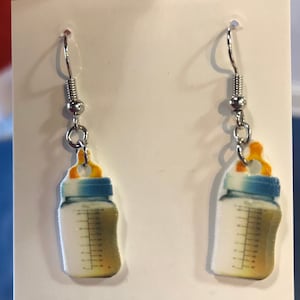 May include: A pair of baby bottle earrings. The earrings feature a silver hook, a small silver bead, and a charm shaped like a baby bottle. The baby bottle charm is white with a blue cap and a yellow nipple. The bottle has measurement markings.