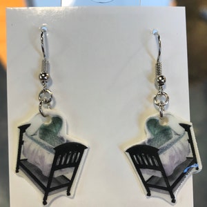 May include: A pair of silver earrings with a baby crib design. The crib is painted in shades of gray, green, and white.