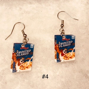 May include: A pair of silver dangle earrings featuring miniature cereal boxes with the Kellogg's Frosted Flakes logo and a cartoon tiger. The boxes are blue, white, and red.