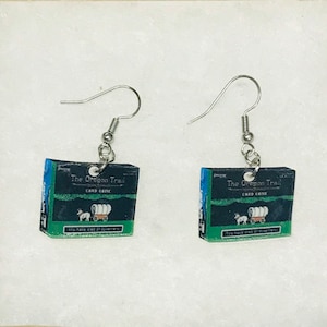 May include: A pair of silver earrings featuring a miniature box of the "The Oregon Trail" card game. The box is green, blue and black with a white wagon and oxen.