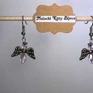 May include: A pair of silver angel wing earrings with pink crystal accents. The earrings are hanging from a wooden dowel rod. The tag reads "Malachi Kitty Bijoux".