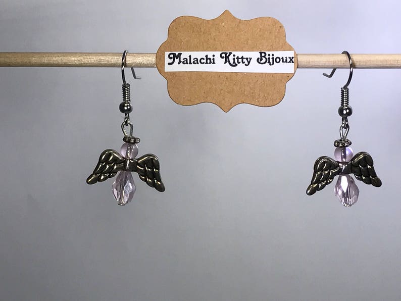 May include: A pair of silver angel wing earrings with pink crystal accents. The earrings are hanging from a wooden dowel rod. The tag reads "Malachi Kitty Bijoux".