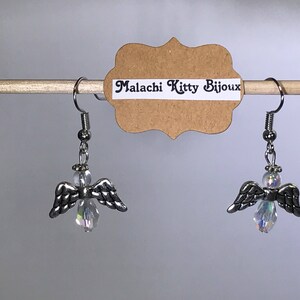 May include: A pair of silver angel wing earrings with clear crystal accents. The earrings are hanging from a wooden dowel rod with a brown paper tag that reads "Malachi Kitty Bijoux".