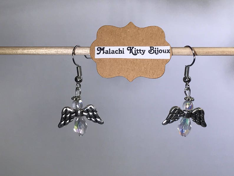 May include: A pair of silver angel wing earrings with clear crystal accents. The earrings are hanging from a wooden dowel rod with a brown paper tag that reads "Malachi Kitty Bijoux".