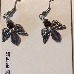 May include: A pair of silver angel earrings with purple crystal accents and brown beads. The earrings are on a white card with the text "Fashion Jewellery" and "Malachi Kitty Bijoux".