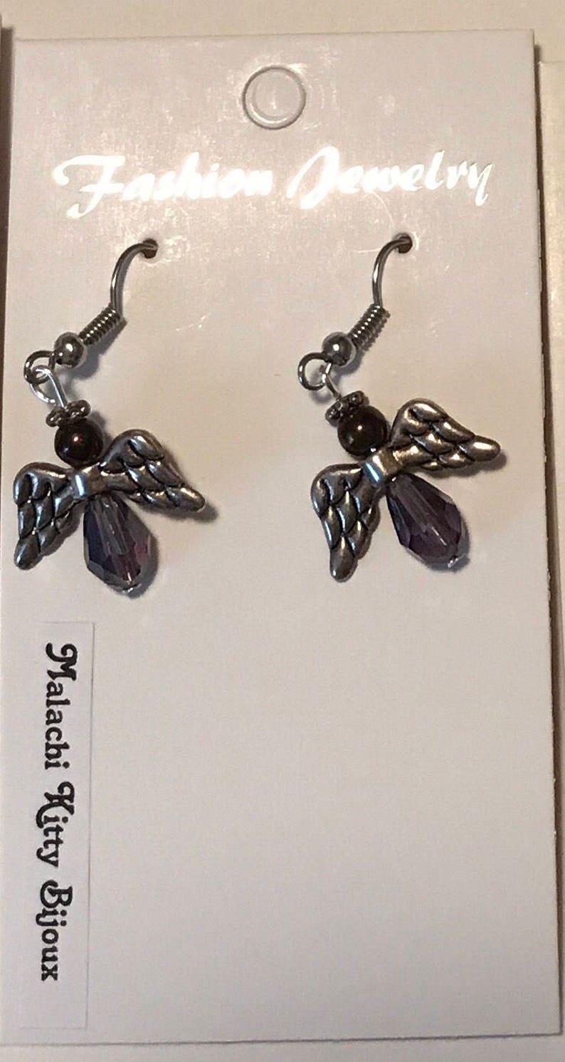May include: A pair of silver angel earrings with purple crystal accents and brown beads. The earrings are on a white card with the text "Fashion Jewellery" and "Malachi Kitty Bijoux".