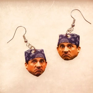 May include: A pair of silver dangle earrings with a blue bandana and a man's face on each earring. The man has a serious expression on his face.
