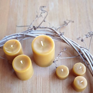 Pu&ograve; includere: Cinque candele di cera d'api gialle di diverse dimensioni, disposte su una superficie di legno con una ghirlanda di vite decorativa. Le candele hanno un aspetto naturale e rustico.