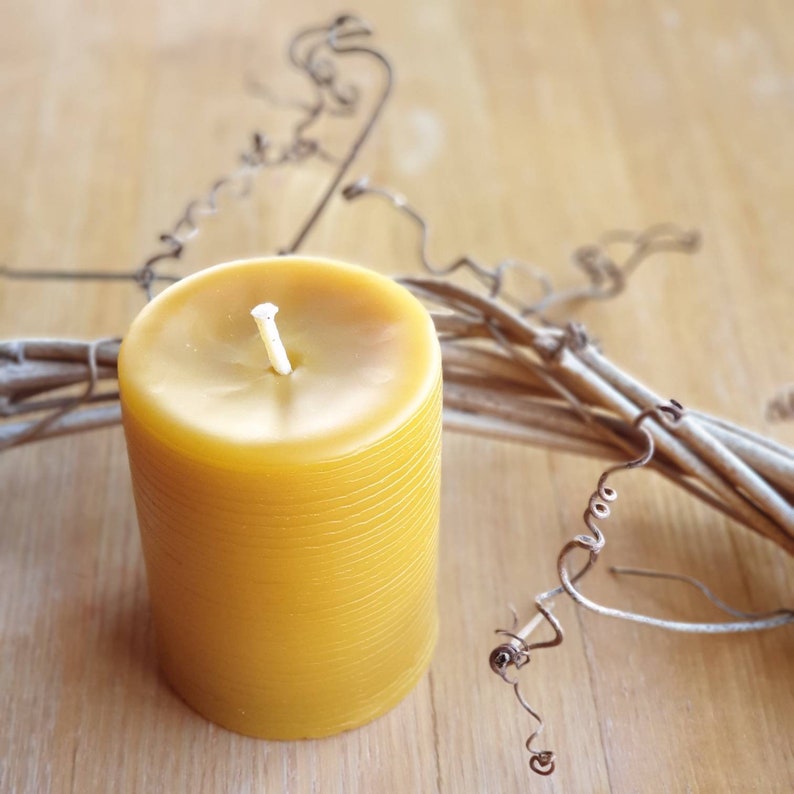 Pu&ograve; includere: Una candela di cera d'api gialla con uno stoppino bianco, appoggiata su una superficie di legno con una ghirlanda di vite decorativa.