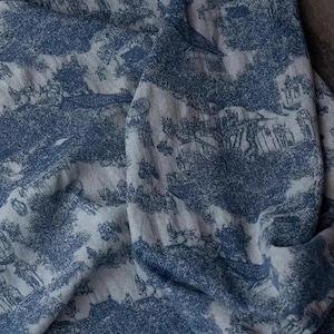 May include: A blue and white fabric with a repeating pattern of a countryside scene. The fabric is folded and draped over a dark surface.