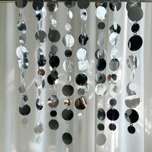 May include: Silver and black round disc garland with a metallic finish. The garland is hanging from a silver rod and is made of multiple strands of discs.