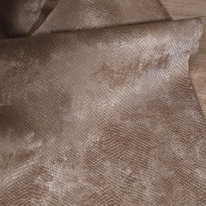 May include: Close-up of a textured, brown fabric with a snakeskin pattern. The material is folded over, revealing the underside. The fabric has a subtle sheen, and the pattern is detailed.