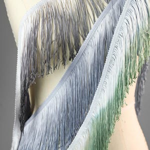 May include: Close-up of a decorative fringe trim with a gradient effect. The fringe transitions from white to grey, then to green. The trim is attached to a white woven base, ideal for embellishing clothing or home decor projects.
