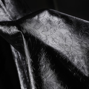 May include: A close-up of black fabric with an embossed floral design. The pattern includes detailed botanical elements, such as leaves and flowers, creating a textured surface. The fabric is folded, showing depth and shadow.