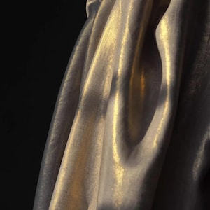 Könnte beinhalten: Nahaufnahme eines drapierten Stoffes mit schimmerndem, goldbeige Aussehen. Das Material ist gefaltet, wodurch Schatten und Glanzlichter entstehen, die seine Textur und seinen Glanz hervorheben. Der Hintergrund ist tiefschwarz und kontrastiert mit dem leuchtenden Stoff.