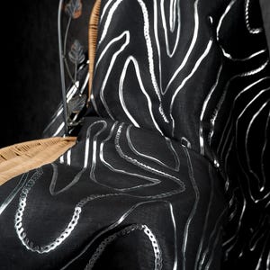 Puede incluir: Tela negra con un diseño plateado abstracto y fluido. La tela está drapeada sobre una silla con un asiento tejido y un respaldo metálico decorativo con detalles de hojas. La estética general es elegante y artística.
