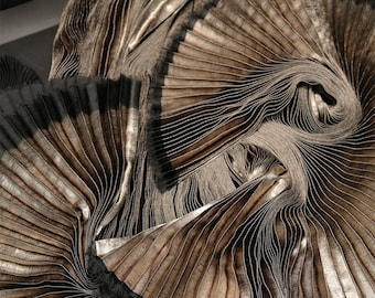 May include: Close-up of a metallic fabric with a pleated texture. The fabric is folded and layered, creating a swirling pattern of light and dark brown hues.